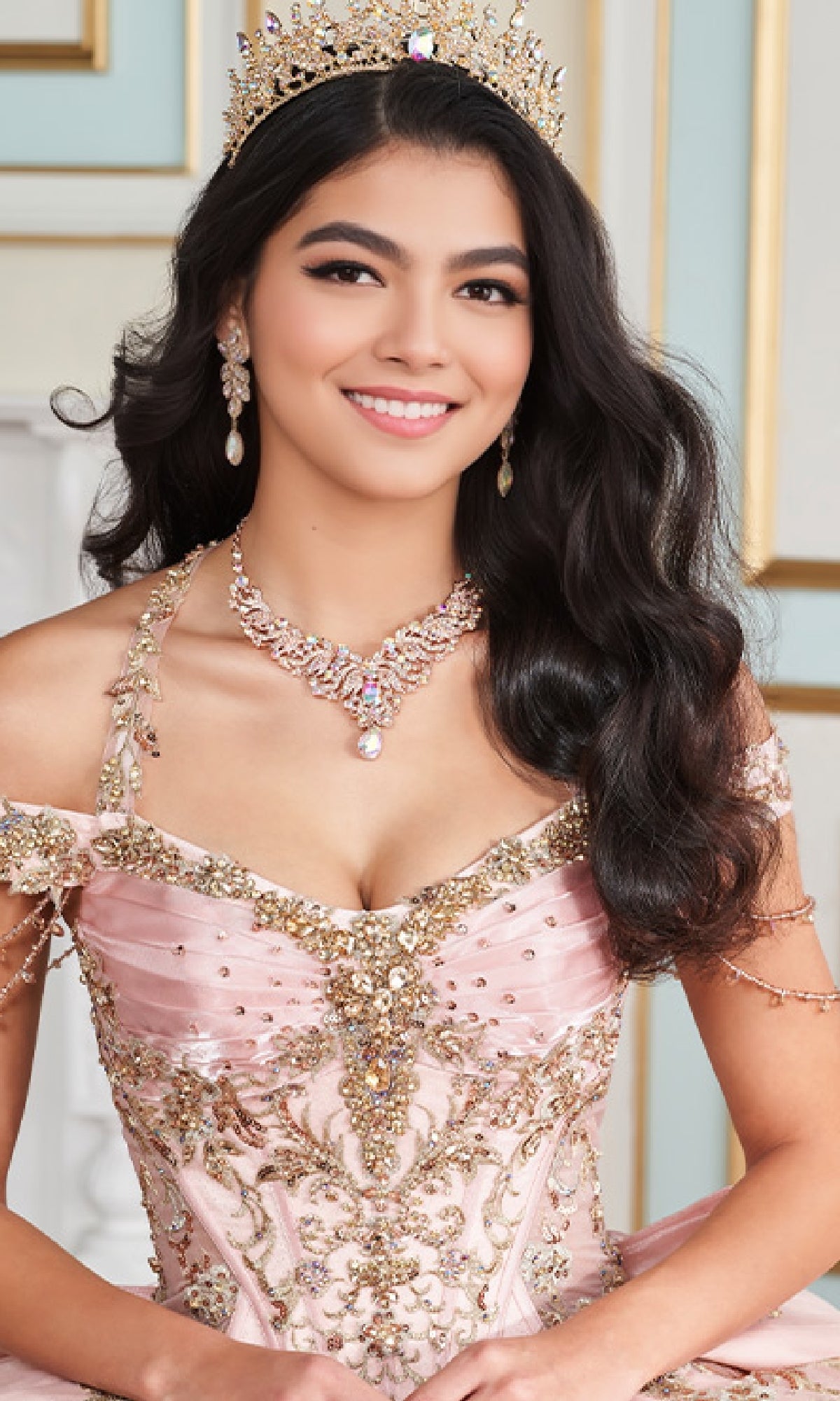 A young woman smiles as she sits wearing the Quinceanera Dress PR30176 By Princesa, a jeweled pink ball gown with metallic beading, a tiara, and matching necklace and earrings, set against an elegant light background.
